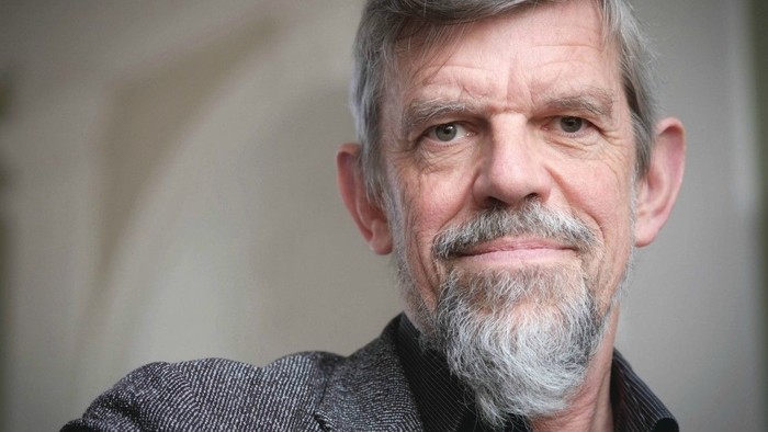 Moden mand med gråt skæg og overskæg poserende eftertænksomt mod en neutral baggrund.