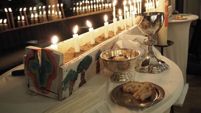 Religiöse Tischdekoration mit Kerzen, Kelch und verzierter Schatulle auf einem weißen Tuch.