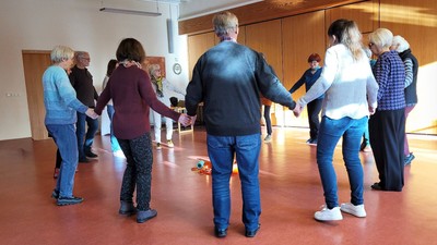 Eine Gruppe von Menschen, die sich an den Händen halten und im Kreis stehen, wahrscheinlich bei einem gesellschaftlichen Tanz oder einer Bewegungsaktivität in Innenräumen.
