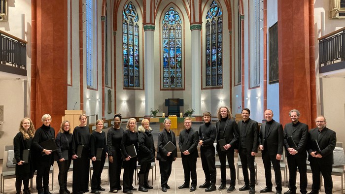 Eine Gruppe von Musikern in formeller schwarzer Kleidung posiert in einer prächtigen Kirche mit farbenfrohen Buntglasfenstern.