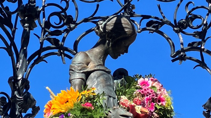 Eine Statue, die leuchtende Blumen unter einem kunstvollen schmiedeeisernen Bogen vor einem klaren blauen Himmel hält.