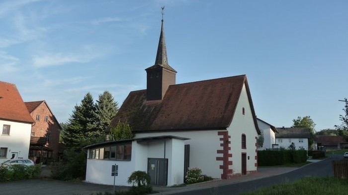 Idyllische Kirche mit rotem Dach und Turm, eingebettet zwischen Wohnhäusern und Grünflächen.