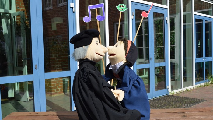 Zwei Puppenprofessoren in Talaren, die flüstern und dabei Musiknoten-Requisiten vor einem Gebäude halten.