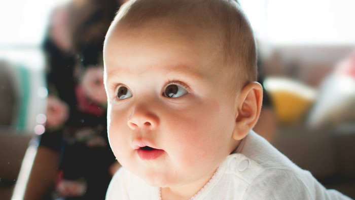 Et spædbarn i en hvid kjole kravler indendørs, mens en smilende voksen kigger på i baggrunden.