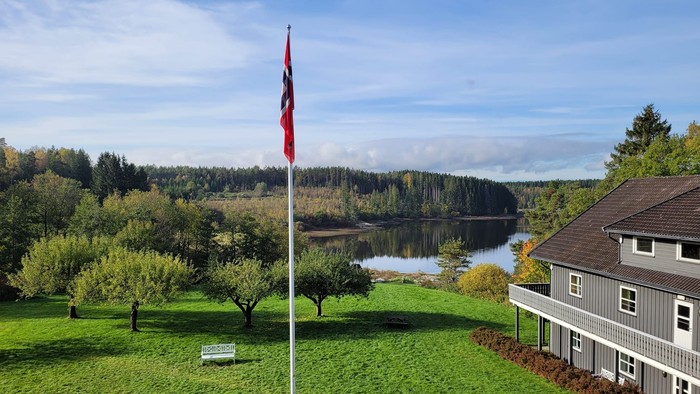 Malerisches Haus am See mit Fahnenmast, üppigem Grün und bewaldeten Hügeln im Hintergrund.
