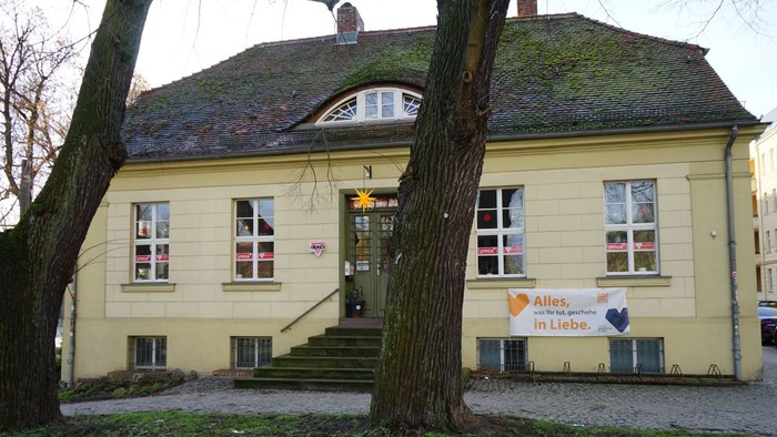 Altbau mit Satteldach und mehreren Fenstern, mittig eine Treppe zur Tür; Davor große Bäume.