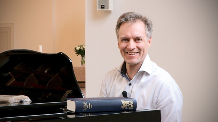 En smilende mand står ved siden af et flygel med en åben nodebog foran sig.