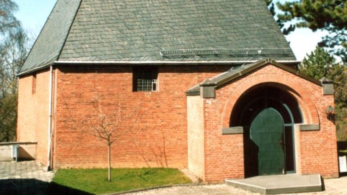 Idyllische Backsteinkapelle mit steilem dreieckigem Dach und markantem Kreuz an der Spitze.