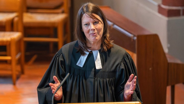 Ein Richter in schwarzer Robe spricht an einem Podium in einem Gerichtssaal mit hölzernen Kirchenbänken und Säulen.