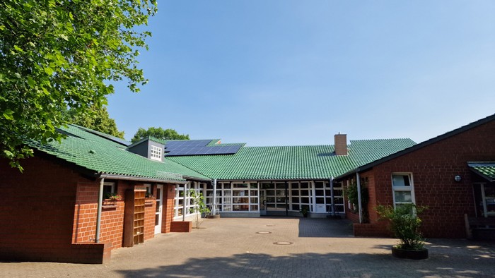 Das Bild zeigt ein rotes Backsteingebäude mit grünem Dach und Solarpanels, umgeben von Bäumen und einem gepflasterten Hof.