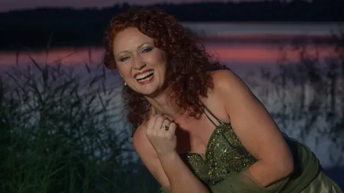 Kvinde med krøllet rødt hår, der griner glædeligt i en grøn kjole ved en malerisk havnefront i skumringen.