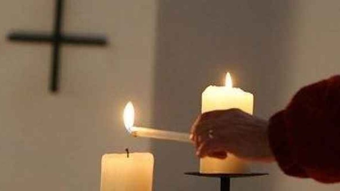 En hånd tænder lys på en stearinlysstøbe foran et lyssted med kors.