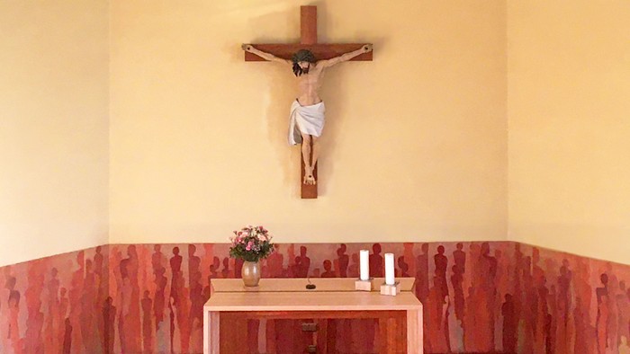 Eine ruhige Kapelle mit einem Altar, einem Kruzifix, einem schlichten Holztisch, Blumen und einer Wandkunst mit roten Silhouetten menschlicher Figuren.