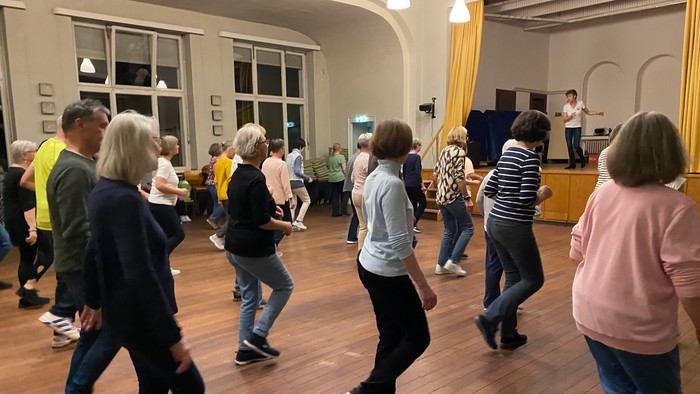 Eine Gruppe von Menschen, die in einem geräumigen, hellen Raum einen Gesellschaftstanzkurs genießt.