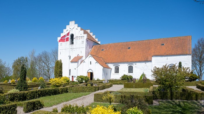 En hvid kirke med rødt tag og dansk flag på tårnet, omgivet af en have med grønne planter og træer