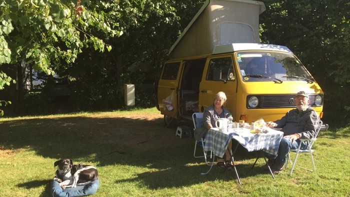 Zwei Personen sitzen in Liegestühlen neben einem gelben Transporter, während ein Hund auf dem Rasen liegt.