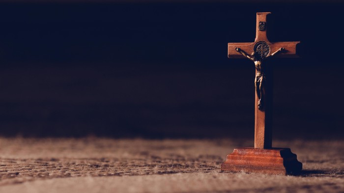 Ein hölzernes Kruzifix mit einer geschnitzten Figur steht auf einer strukturierten Oberfläche vor dunklem Hintergrund.