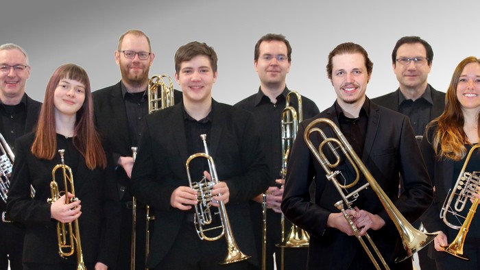 Musikerinnen und Musiker eines Blechbläserensembles stehen nebeneinander vor hellem Hintergrund. Sie tragen schwarze Konzertkleidung und halten verschiedene Blechblasinstrumente wie Trompeten und Posaunen in den Händen. Alle blicken in die Kamera und läch