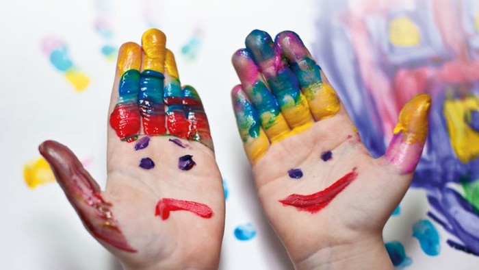Zwei Hände mit bemalten Gesichtern und bunten Fingern – die eine mit finsterer Miene, die andere lächelnd – mit einer Spielzeugeisenbahn im Hintergrund.