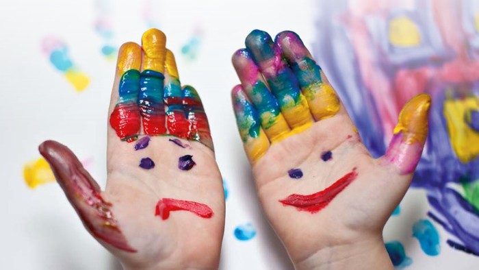 Zwei Hände mit bemalten Gesichtern und bunten Fingern – die eine mit finsterer Miene, die andere lächelnd – mit einer Spielzeugeisenbahn im Hintergrund.