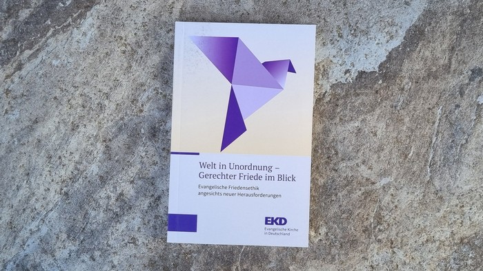 Eine Broschüre mit dem Titel *„Welt in Unordnung“*, angebracht an einer strukturierten Steinwand mit EKD-Logo.