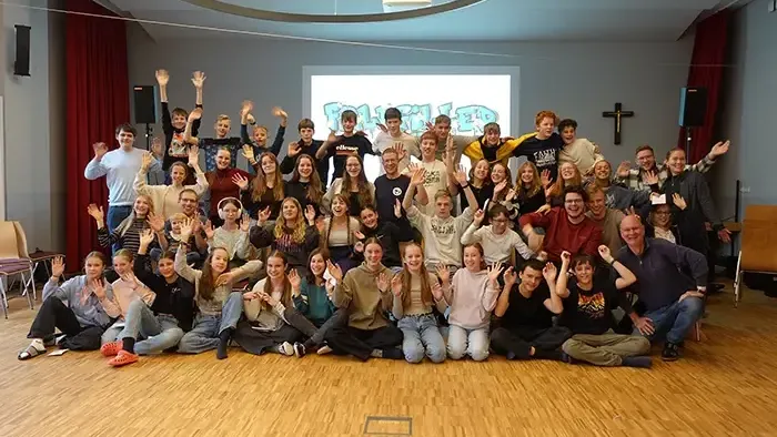 Eine große Gruppe von Menschen posiert fröhlich in einem Raum mit Holzboden und Kirchenstuhl.