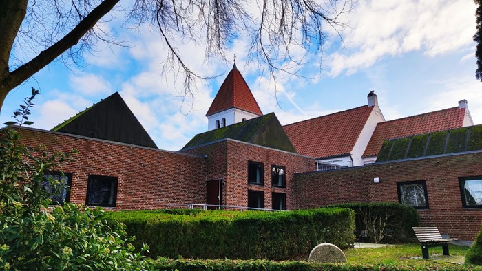 Et rødt teglstenshus med spiret og omgivet af grønne buske og en træ.