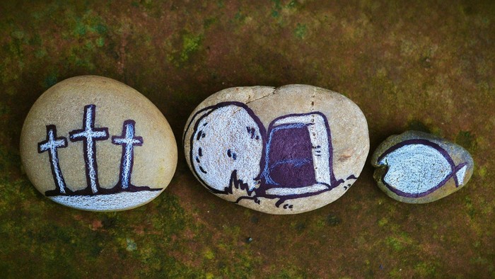 Bemalte Steine zeigen Kreuze, eine offene Bibel und ein Augensymbol auf moosigem Boden.