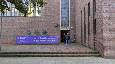 Person betritt ein Backsteingebäude mit dem Schild *„Herzlich willkommen in der Matthäikirche!“*.