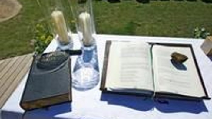 Auf einem Tisch im Freien stehen eine Flasche Wasser, ein Glas und ein aufgeschlagenes Buch.