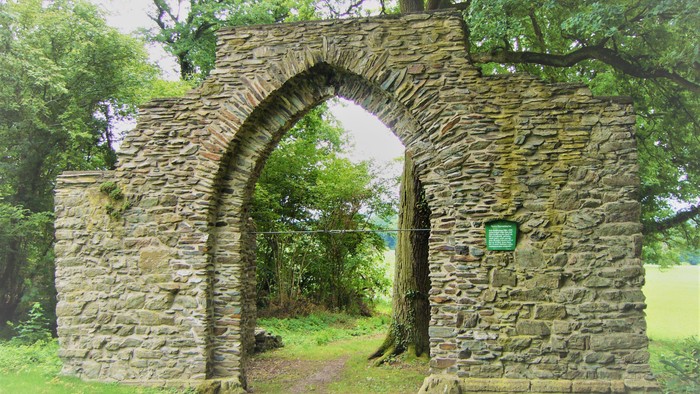Ein alter Steinbogen steht in einem Park umgeben von Bäumen und Gras.