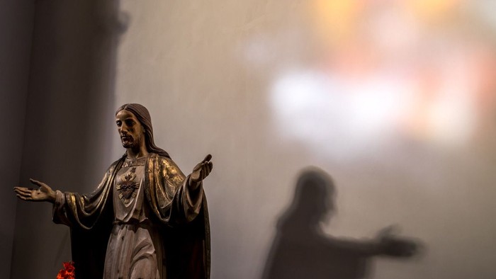 Eine Statue der Jungfrau Maria mit ausgebreiteten Armen in einer Kirche, im Hintergrund eine verschwommene Gestalt.