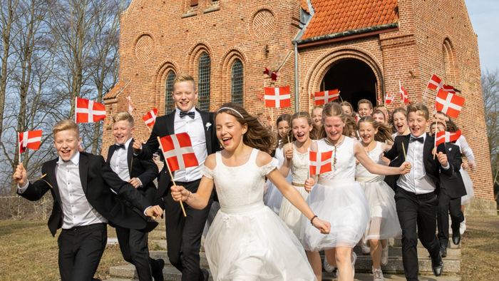 En glad gruppe fejrer uden for en murstenskirke, mens de vinker med dannebrog og er klædt i festligt tøj.