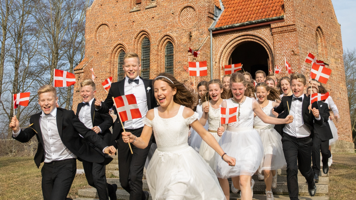 En glad gruppe fejrer uden for en murstenskirke, mens de vinker med dannebrog og er klædt i festligt tøj.