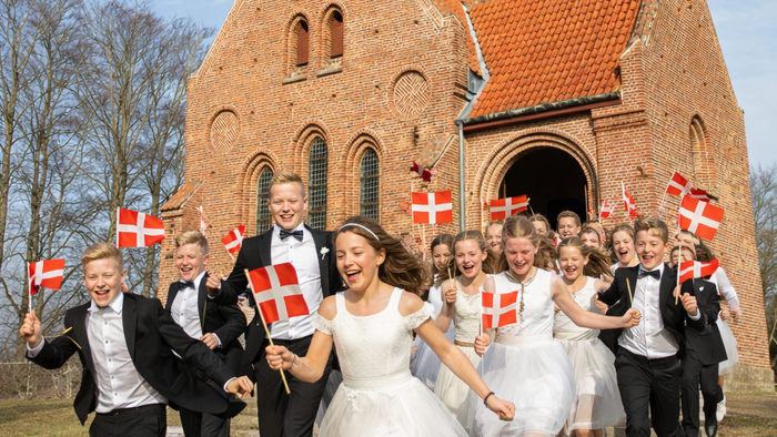 En glad gruppe fejrer uden for en murstenskirke, mens de vinker med dannebrog og er klædt i festligt tøj.