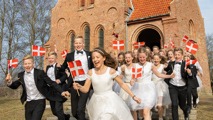 En glad gruppe fejrer uden for en murstenskirke, mens de vinker med dannebrog og er klædt i festligt tøj.