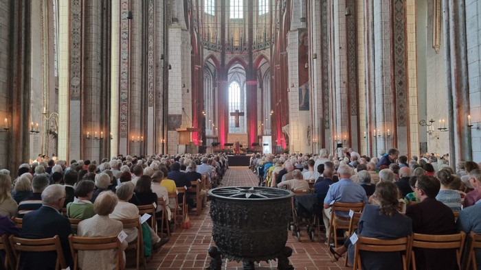 Große Menschenmenge in einer Kathedrale, viele sitzen auf Stühlen, hohe Decken, bunte Fenster.