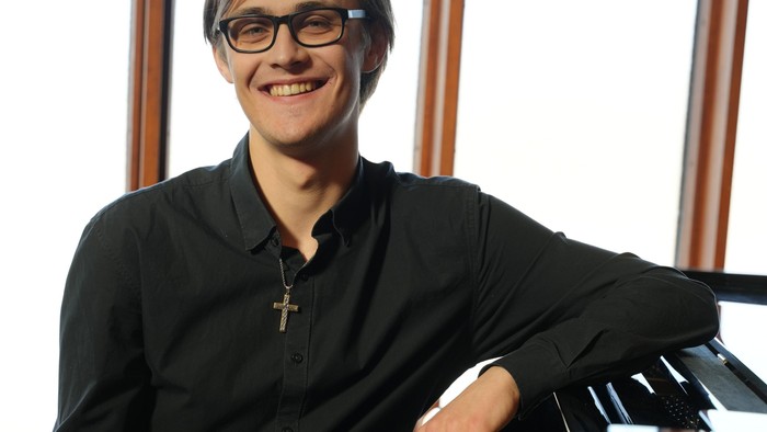 En smilende mand med briller sidder ved siden af et flygel iført en sort skjorte.