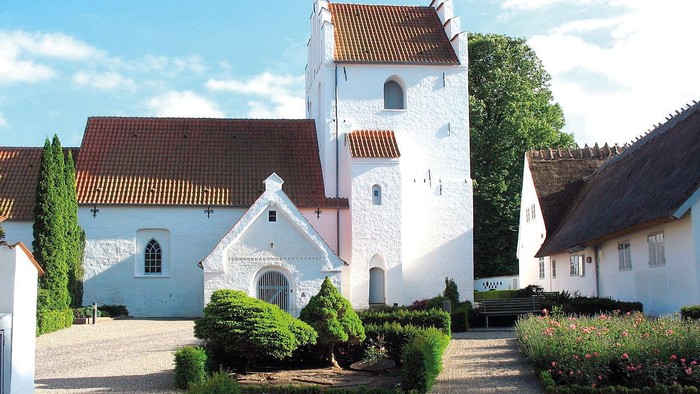 En smuk og hvid kirke med tårn og røde tag i en trædit og grønne omgivelser