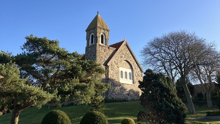 Hyggelig stenkirke med klokketårn omgivet af veltrimmet grønne områder under en klar blå himmel.