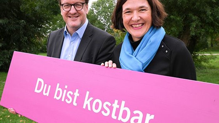 Ein Mann und eine Frau halten vor grüner Kulisse ein pinkfarbenes großes Schild mit der Aufschrift 
