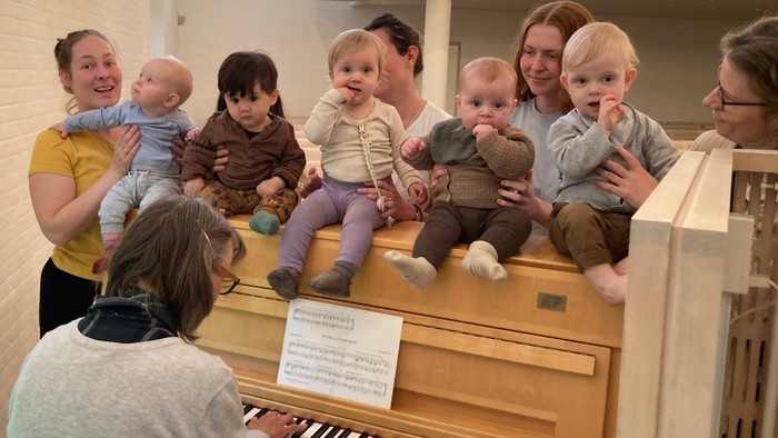 En gruppe børn og voksne sidder på et piano i en lokal.