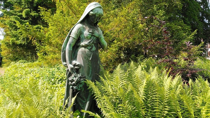 Bronzestatue einer trauernden Frau, die eine Hand an die Brust legt und mit der anderen Blumen hält