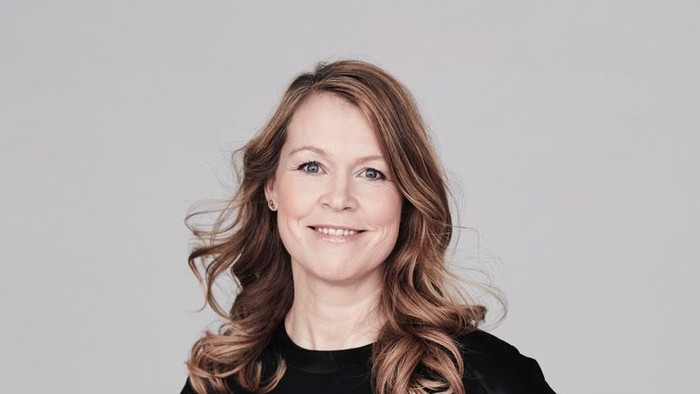 En kvinde med krøllet hår smiler selvsikkert i en elegant sort top på en neutral baggrund.