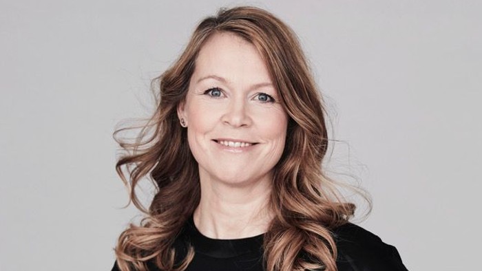 En kvinde med krøllet hår smiler selvsikkert i en elegant sort top på en neutral baggrund.