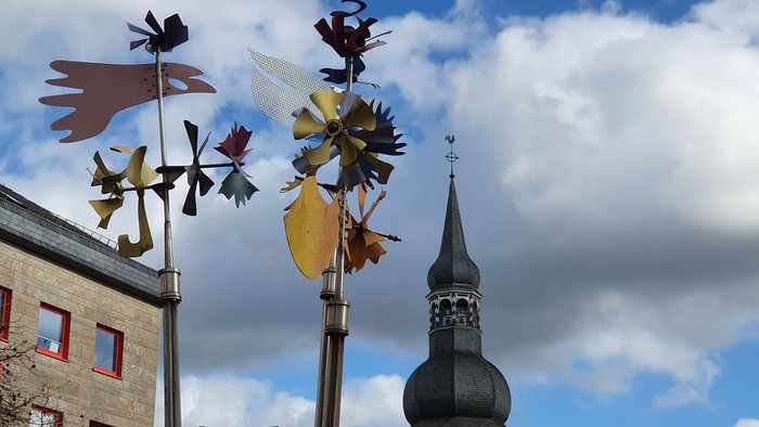 Bunte Windräder an einem Pfahl neben einem historischen Backsteingebäude und einem Kirchturm.