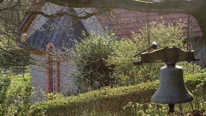 Eine ländliche Gartenszene mit einer hängenden Glocke und üppigem Grün, das die bunten Dachziegel eines Hauses widerspiegelt.