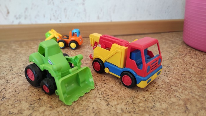 Helle Spielzeug-Baufahrzeuge, darunter ein grüner Bulldozer, ein bunter Abschleppwagen und ein kleiner gelber Bagger auf einer Korkoberfläche.