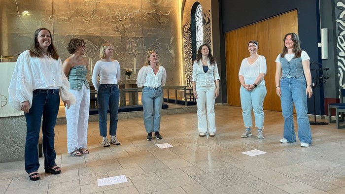 En gruppe på syv kvinder står på række med afstandsmarkører på et poleret gulv indendørs.