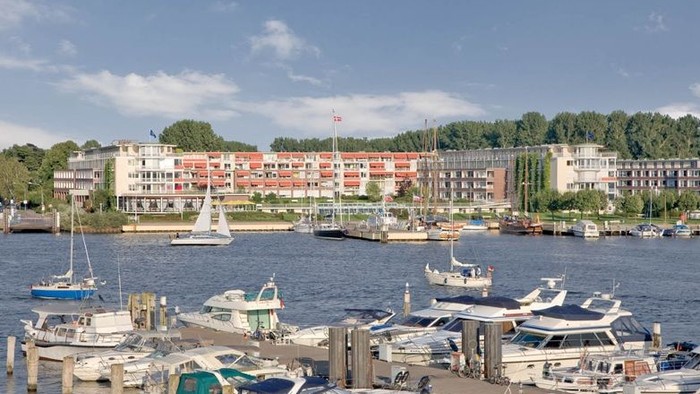 Eine ruhige Marina, gefüllt mit Booten, gesäumt von bunten Gebäuden am Wasser und Bäumen unter einem klaren Himmel.